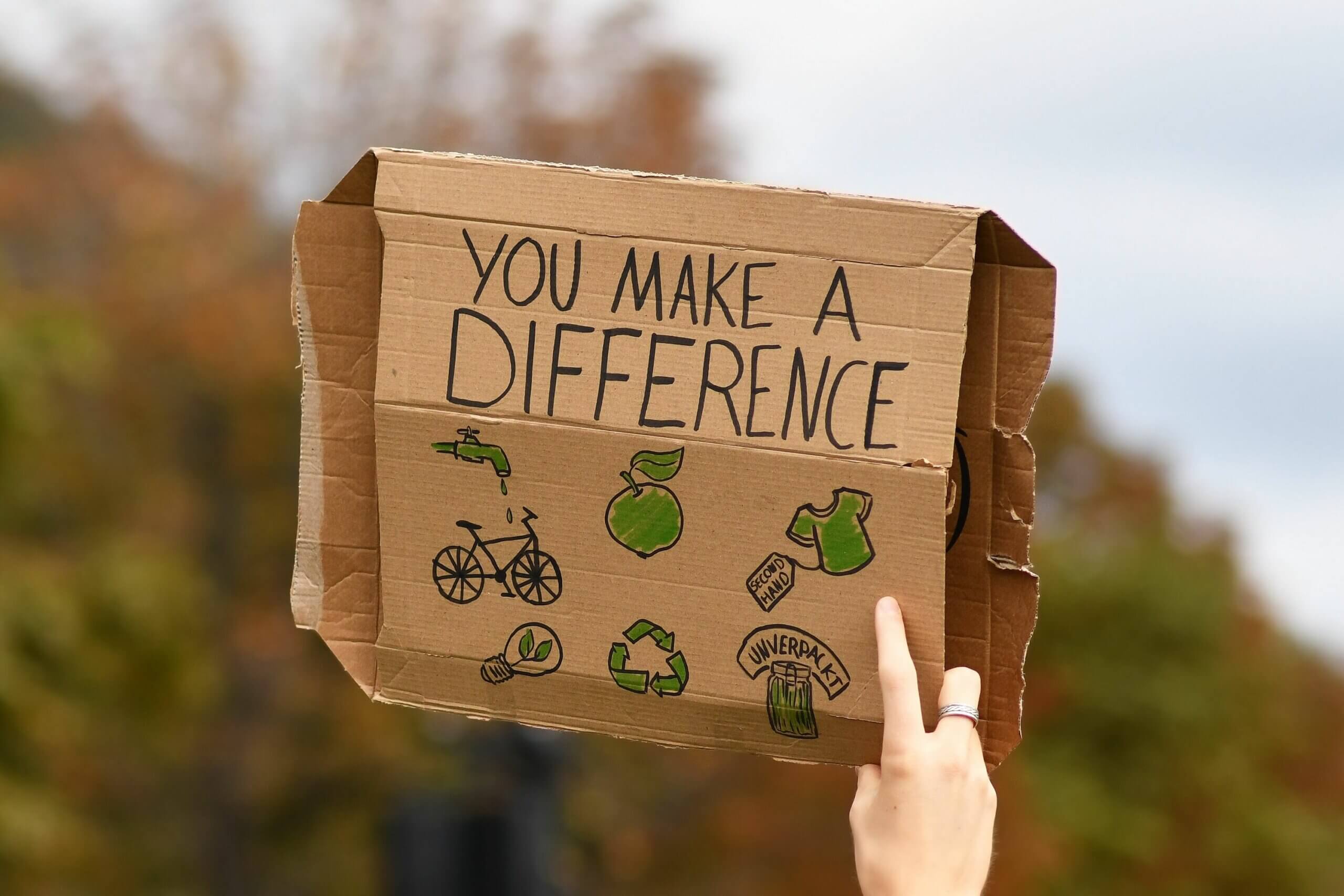 Ein bemaltes Pappschild wird in die Luft gehalten. Auf dem Schild steht: You make a difference. Auf dem Schild sieht man zudem aufgemalte Symbole: ein tropfender Wasserhahn, ein Apfel, ein T-Shirt mit der Aufschrift second hand, das Kreislauf-Zeichen, eion Glas mit der Aufschrift unverpackt, eine grüne Glühbirne und ein Fahrrad.||||