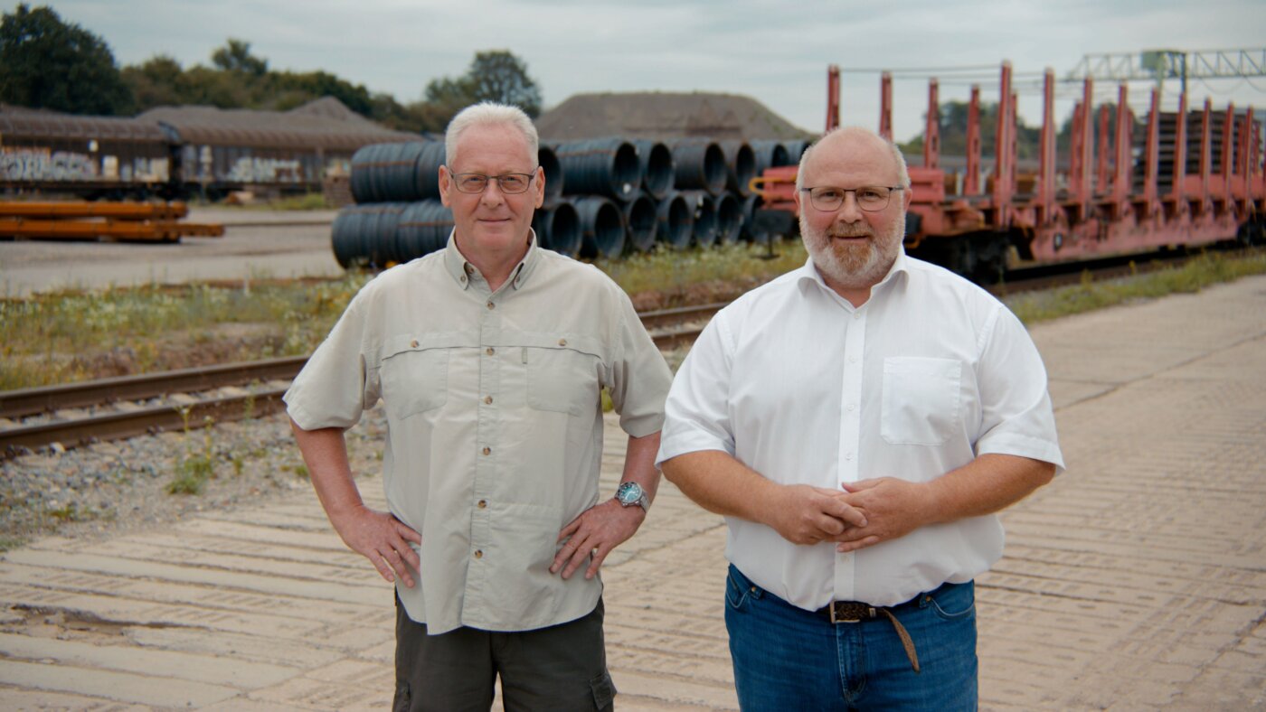 Auf dem Foto sieht man zwei ältere Personen, im Hintergrund ein Bahn-Industriegelände mit Rohren und Sandbergen.